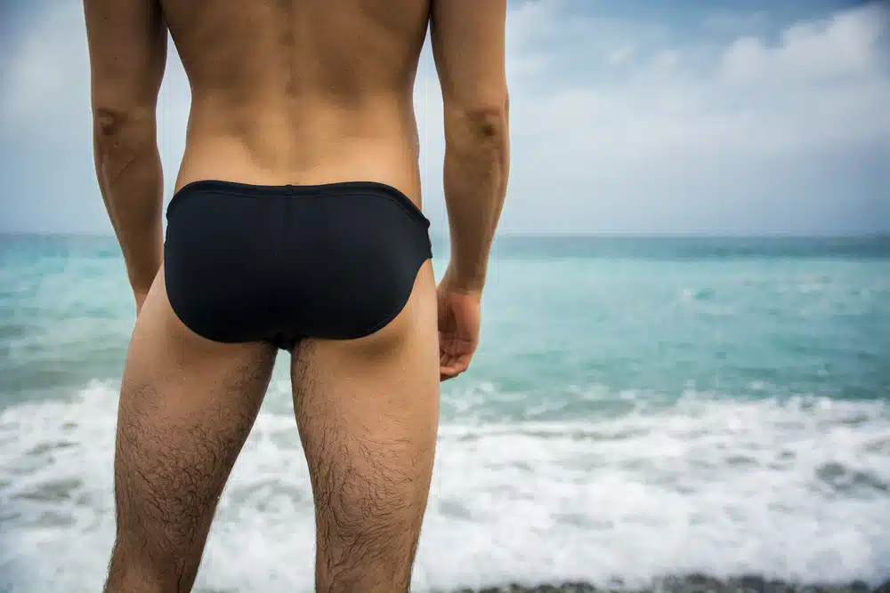 Man in swimwear standing by ocean showing results of gluteal enhancement with improved projection and balance.