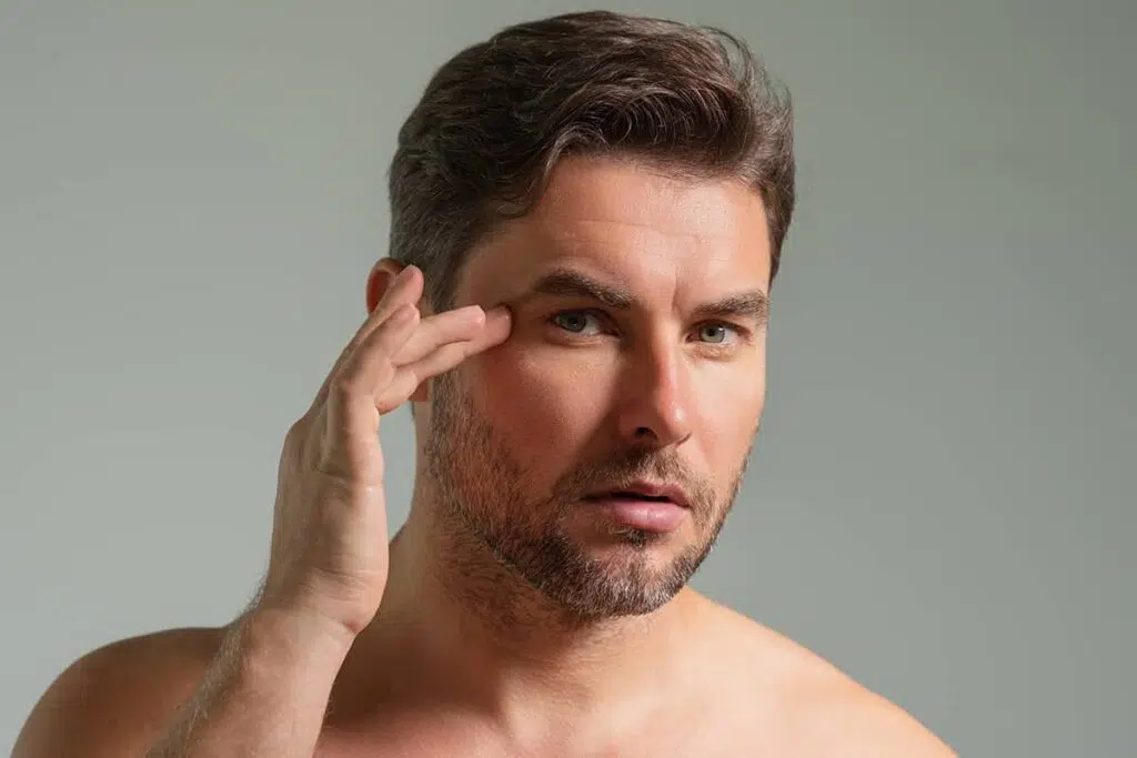 A man with a well-groomed beard examining his facial contours in the mirror, touching the skin near his eye to check for signs of aging.