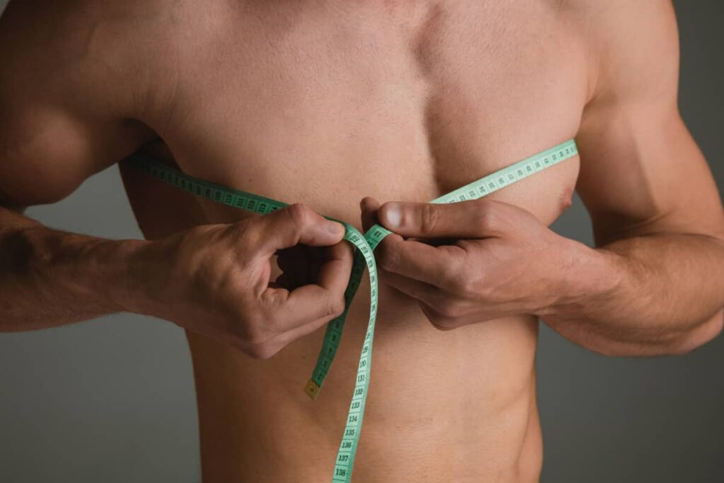 A muscular man using a green measuring tape to measure the circumference of his chest, highlighting pectoral development and symmetry.